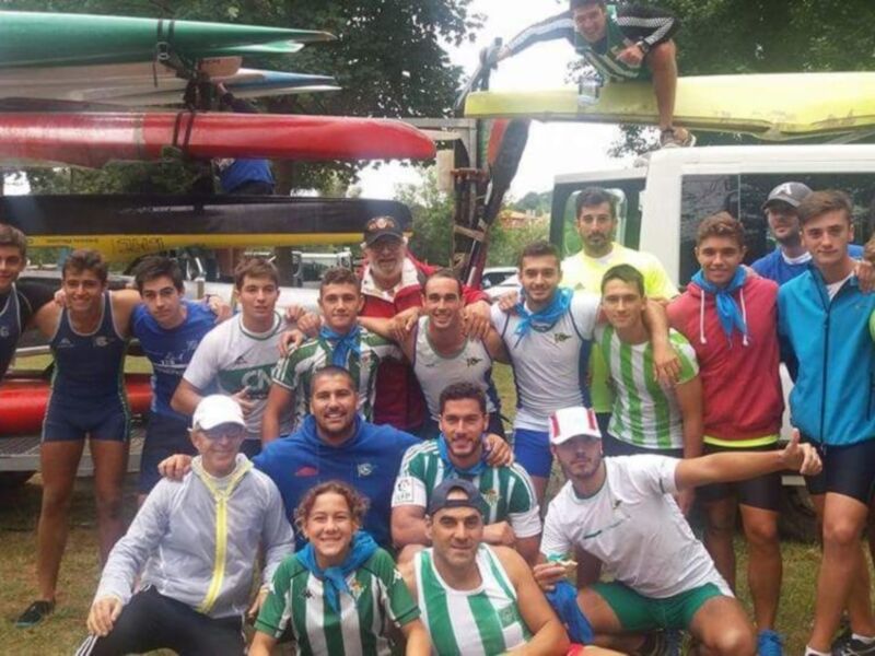 Un grupo de jóvenes deportistas posa en un campo verde, con remos y canoas apilados detrás de ellos. Algunos llevan camisetas deportivas coloridas, mientras otros usan gorras y chalecos. La escena sugiere un día de entrenamiento o competencia en canoa.