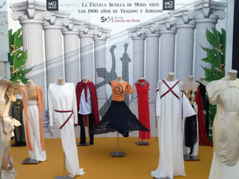 Exposición de moda con vestidos y accesorios en un fondo con columnas romanas y hojas verdes.