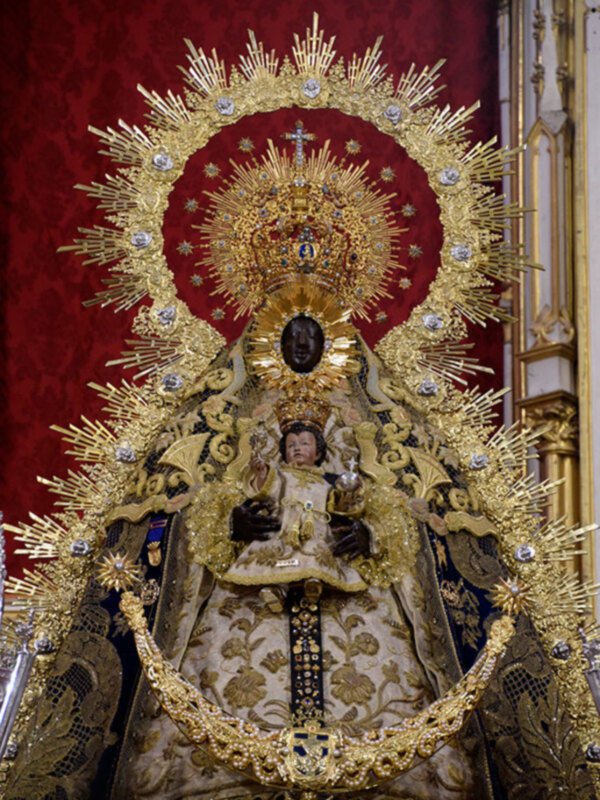 Imagen de la Virgen María con un manto dorado y una corona estrellada. La imagen se encuentra en un entorno religioso, posiblemente una iglesia, con fondos rojos y detalles dorados. La Virgen sostiene a un niño en su regazo, ambos vestidos con vestimentas ornamentadas. La imagen transmite una sensación de veneración y devoción.