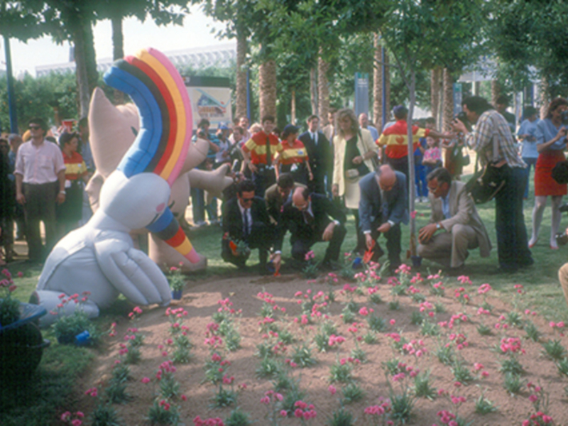 Una imagen de una celebración con globos en forma de arcoíris y un personaje de dibujos animados, rodeada por una granja con flores rosadas y un grupo de personas en una carpa.