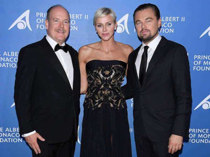 Tres personas elegantes posando juntos en una alfombra roja con fondo azul y logotipos de la Princesa Grace Foundation.