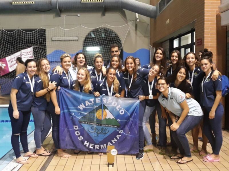 Equipo femenino de waterpolo posando con una bandera de la ciudad hermana en un gimnasio.