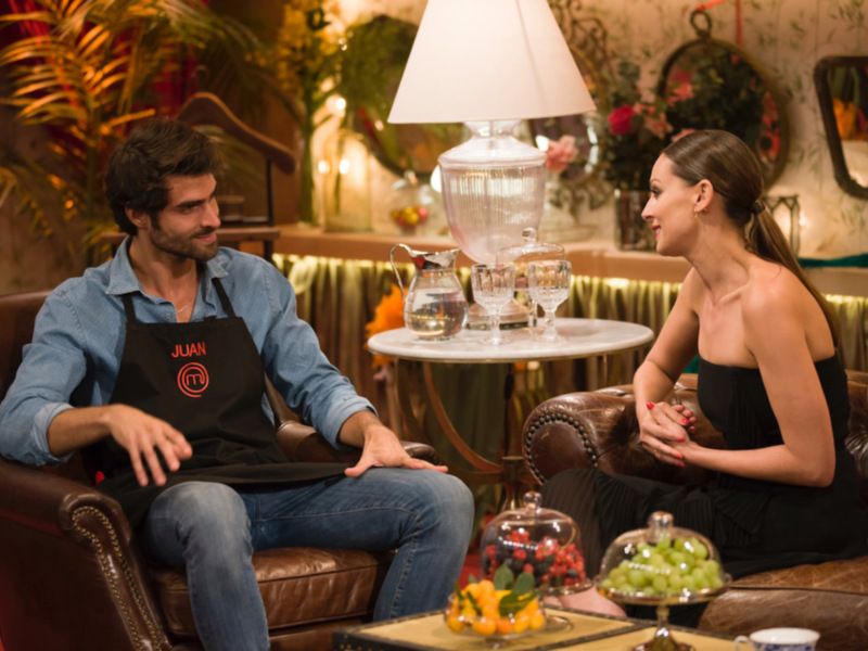 Un hombre y una mujer charlan en un ambiente elegante con flores, una lámpara y una mesa de cristal.