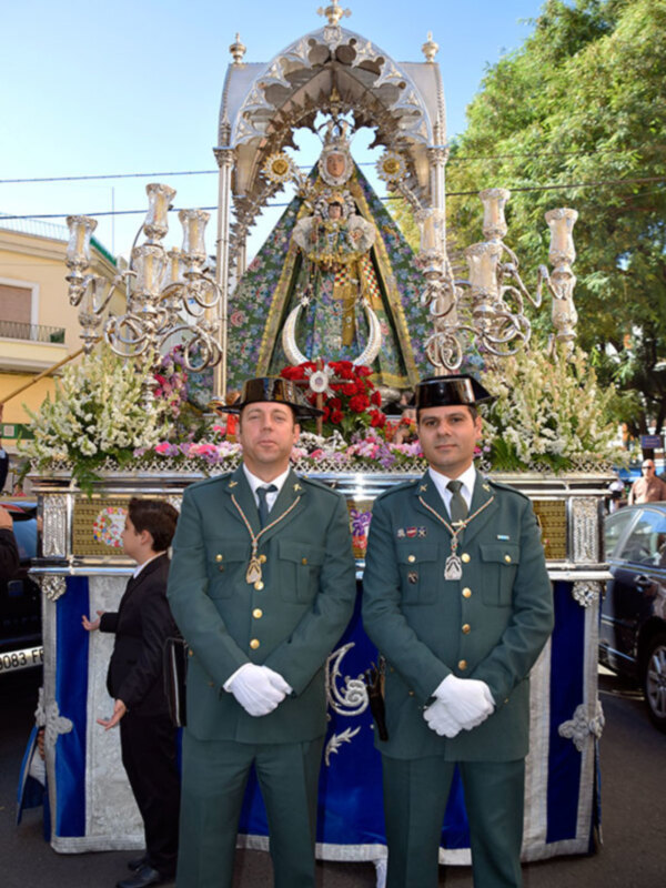 Dos hombres en uniforme militar posan frente a una estatua religiosa durante una procesión festiva. El arco central está adornado con flores y decoraciones, y los hombres llevan insignias militares y sombreros militares.