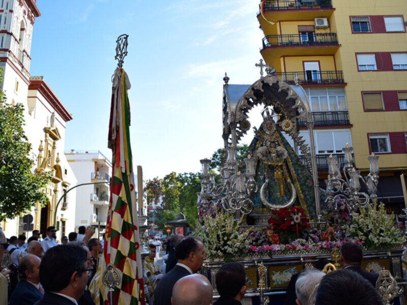 Procesión religiosa con imagen de la Virgen María en un carro ornado con flores y candelabros, acompañada por fieles y banderas.