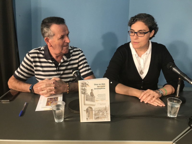 Dos personas sentadas en una mesa con un libro abierto en frente de ellos, uno de ellos sostiene un microfono.