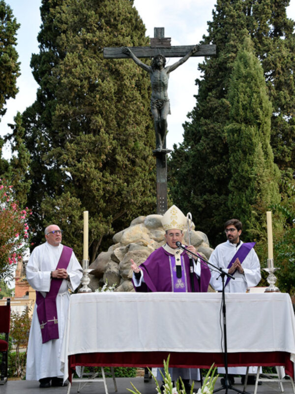 Crucifijo en un entorno religioso con tres personas vestidas con vestimentas sacerdotales. En el fondo, hay árboles y una pila de piedras. En el centro, hay un altar con velas y un microfono.