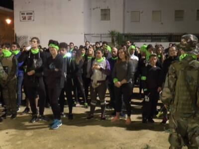 Una multitud de personas con gorros verdes y cintas reflectantes se concentra en una calle nocturna. En el fondo, un edificio con ventanas y carteles se destaca. Dos individuos vestidos de uniforme militar observan la escena desde el lado izquierdo.