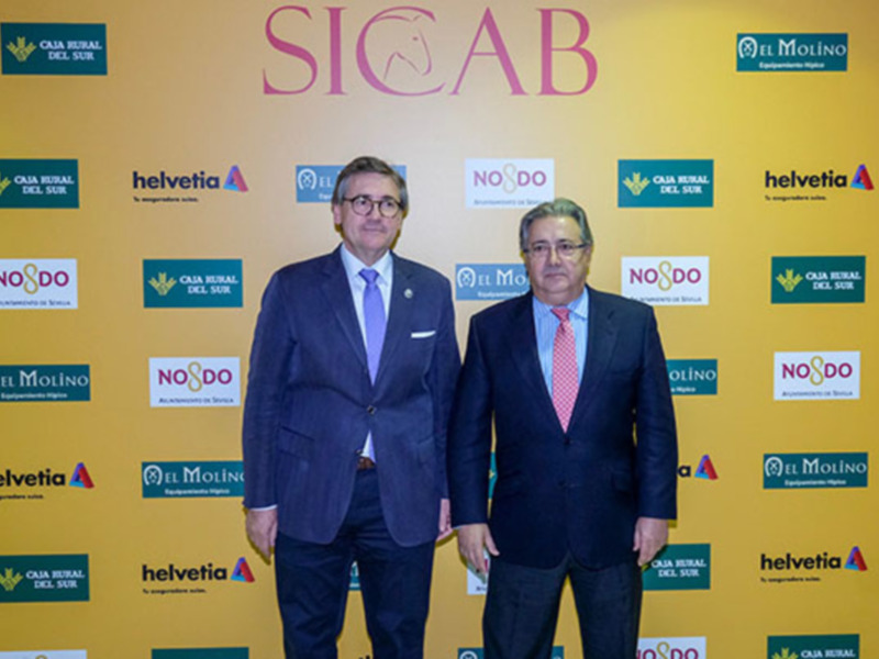 Dos hombres en trajes de negocios posan frente a un fondo con logos de empresas.