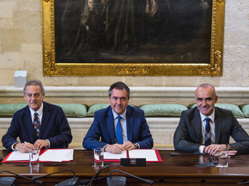 Tres hombres en trajes sentados frente a una mesa con documentos. Detrás de ellos, un lienzo enmarcado grande sobre una pared de ladrillo. La escena parece ser formal y probablemente se refiere a un evento o reunión importante.