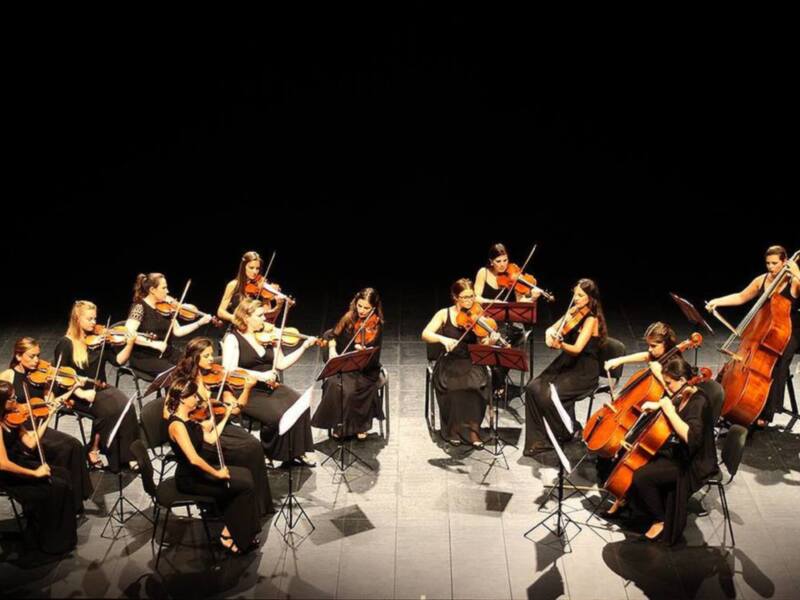 Orquesta femenina en concierto, interpretando música clásica en un escenario oscuro.