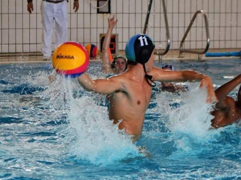Un niño juega al waterpolo en una piscina de agua azul, con un balón rojo y amarillo. El niño lleva un casco azul y blanco, mientras otros jugadores intentan alcanzar el balón. La pared de la piscina es de azulejos blancos y grises, con un barandilla metálica en el fondo.