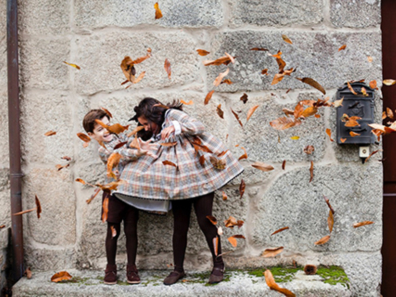 Dos niños disfrutan de la caída de hojas en otoño, frente a una pared de piedra. El ambiente es cálido y relajante, reflejando la belleza de la temporada.