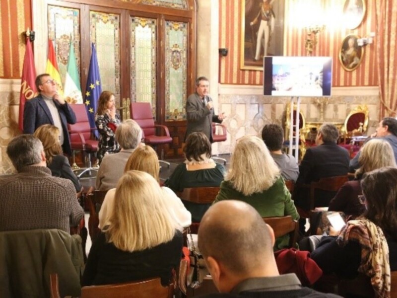 Una sala de conferencias con personas sentadas en sillas, un hombre hablando en el escenario y banderas de España y la UE en el fondo.