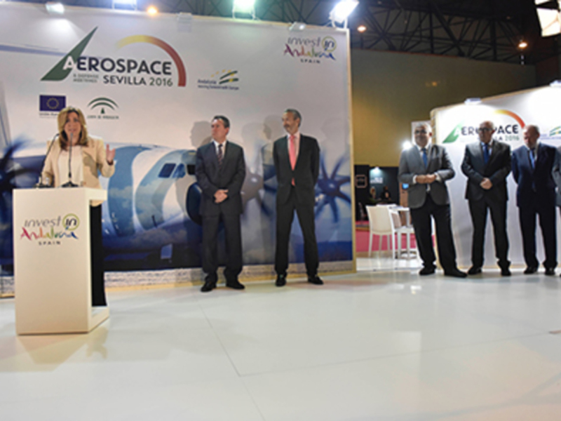 Presentación en el evento AEROSPACE Sevilla 2016 con una bandera española y un avión en el fondo. El evento promueve la inversión en España.