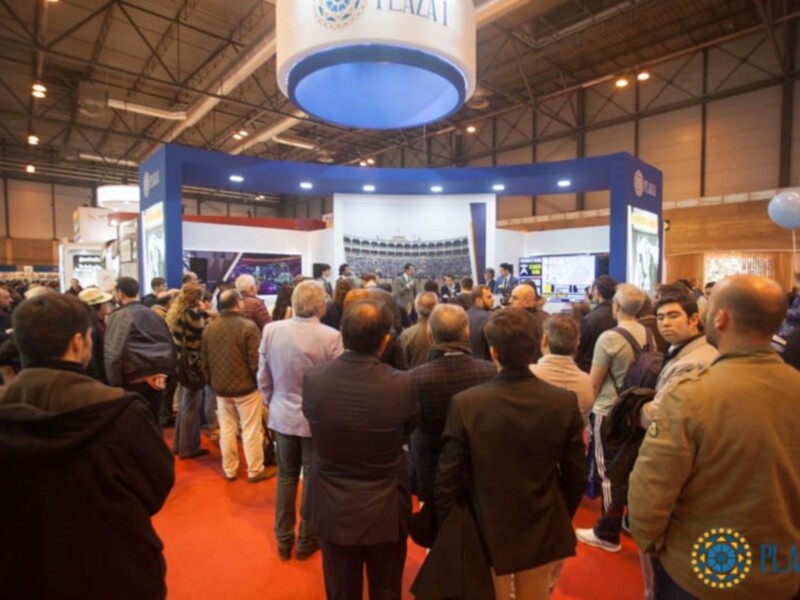 Una multitud de personas en una feria o evento, con stands y pantallas de proyección. La iluminación es intensa, destacando el ambiente de la exposición.