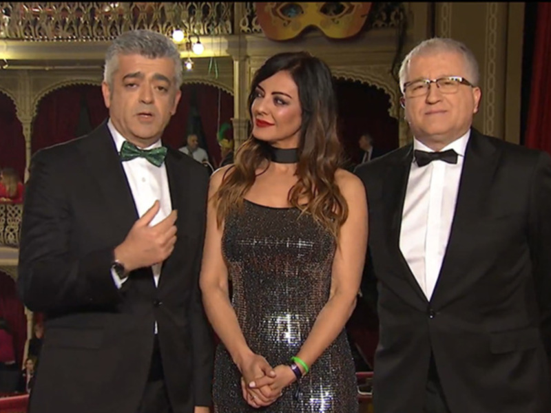 Tres personas elegantes en trajes de fiesta posan juntas en un escenario con decoración teatral.