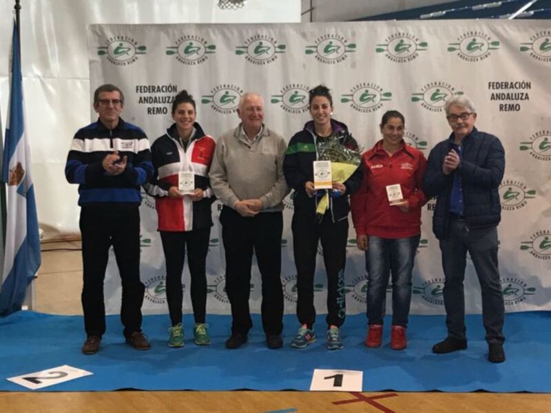 Equipo de atletas celebrando en un podio con medallas, frente a una bandera y un banner de la Federación Andaluza de Atletismo.