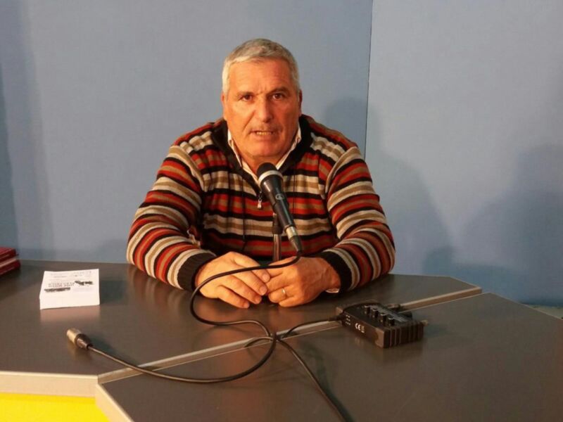 Un hombre con cabello gris y barba, vistiendo una camiseta de rayas, se sienta en un escritorio con un micrófono y cables. La pared de fondo es azul claro, y en el escritorio hay un pequeño sobre y una tarjeta.