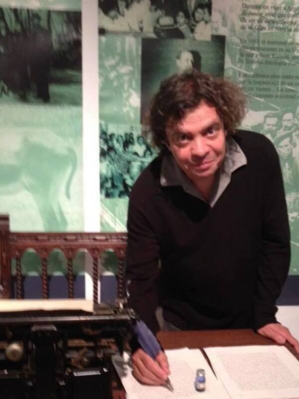 Un hombre con cabello rizado está sentado en frente de una máquina de escribir antigua, escribiendo en un papel. El fondo muestra imágenes históricas y textos sobre el movimiento comunista.