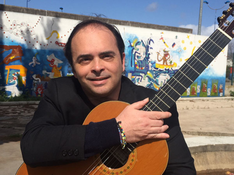 Un hombre sostiene una guitarra acústica frente a un mural colorido.