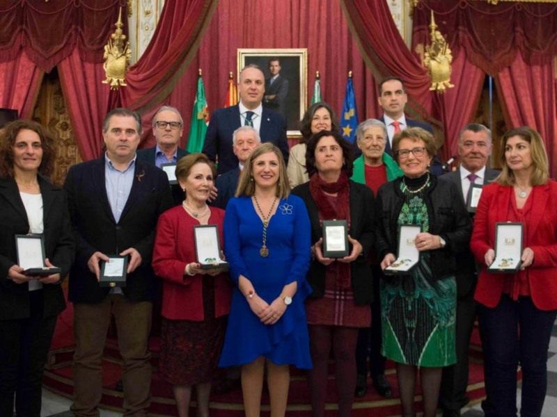 Una imagen de una ceremonia formal donde varias personas sostienen medallas. El fondo es elegante con cortinas rojas y un retrato en el centro. La iluminación es brillante, destacando a los participantes y sus medallas.