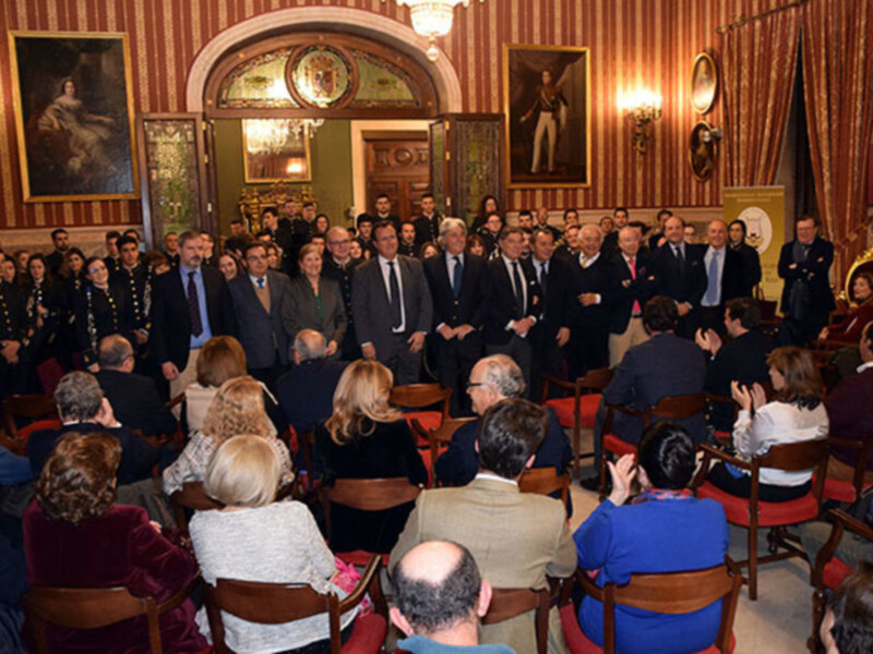 Una sala de audiencias con personas sentadas en sillas, algunos vestidos formales y otros más informales. Paredes decoradas con cuadros, cortinas rojas y una chimenea en el fondo.