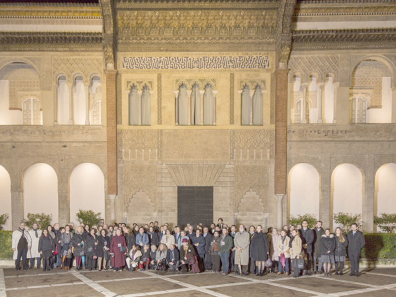 Una imagen de una multitud frente a un edificio histórico con arcos y columnas doradas. La gente está vestida formalmente, posando para una foto colectiva en un entorno urbano.