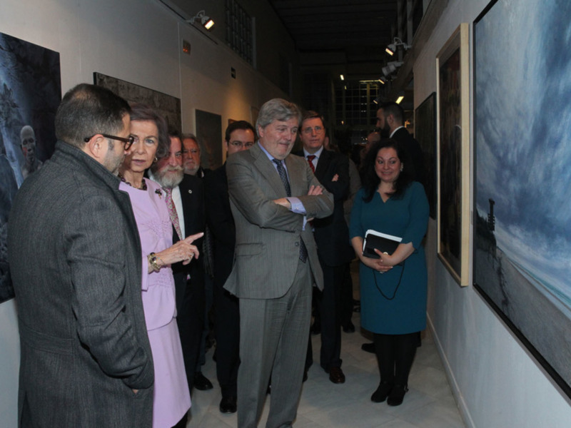 Persona en una exposición de arte, conversando con visitantes mientras examinan obras de arte en la pared.