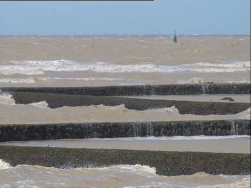 Playa con marea baja, mostrando la intersección entre el agua y la arena, con un faro en el horizonte.