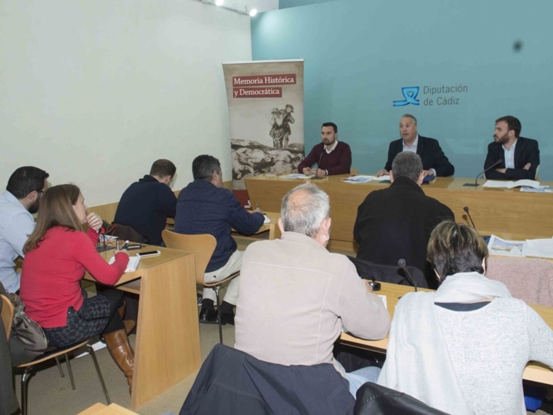 Una reunión formal en un salón de conferencias con personas sentadas en mesas y un panel de tres personas en el frente, con una pancarta que dice 'Memoria Histórica y Democrática' y la logotipo de la Diputación de Cádiz.