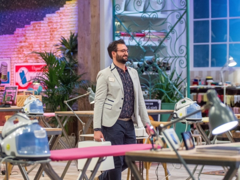Un hombre con una camisa estampada y una chaqueta beige camina por un área con mesas y sillas, con un fondo que incluye una pantalla de televisión con un diseño de pirámides y una iluminación azul.