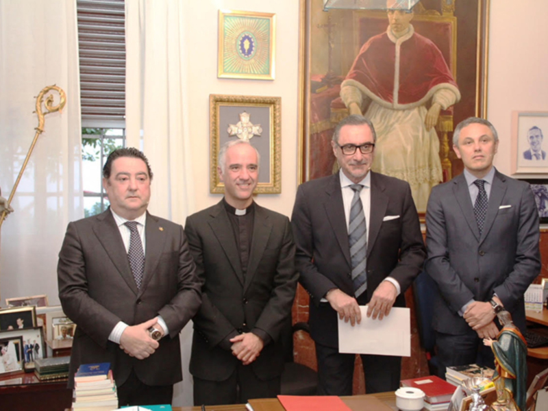 En un entorno formal, cuatro hombres en trajes posan frente a una mesa con documentos y una estatua religiosa. La imagen sugiere un encuentro oficial o negociación.
