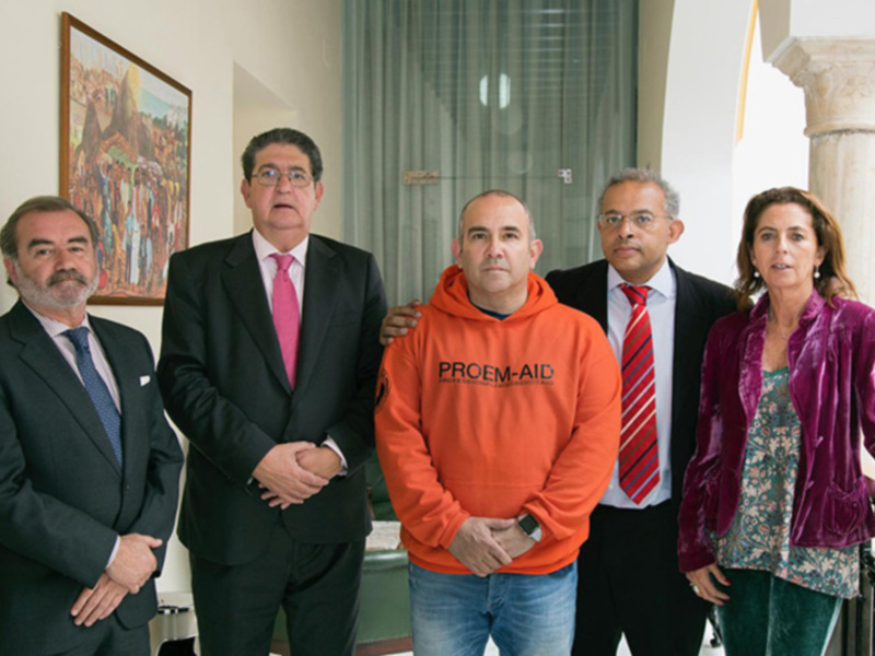 Un grupo de personas, incluyendo un hombre en un suéter naranja con 'PROEM-AID' y otros en trajes, posan juntos en un interior bien iluminado.