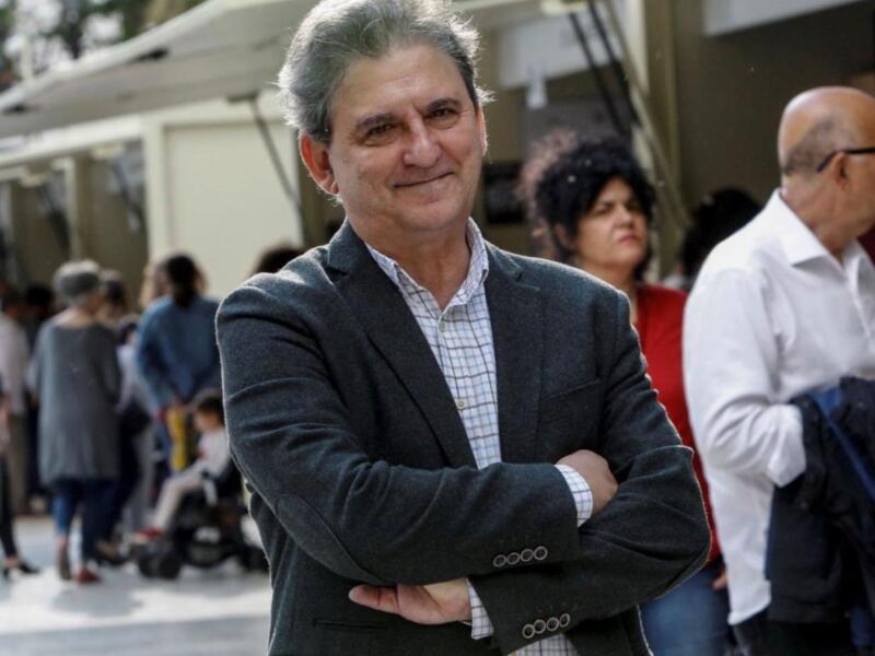 Un hombre con cabello gris y barba, vestido con una chaqueta de traje y camisa blanca, con las manos cruzadas frente a él. La imagen parece ser de un evento público, con personas en el fondo y una estructura blanca al fondo.