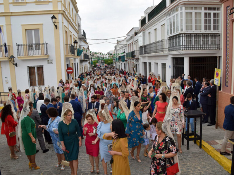 Una multitud de personas vestidas con trajes tradicionales camina por una calle adoquinada, rodeada de edificios blancos y amarillos. La imagen captura una celebración festiva, posiblemente una procesión o fiesta local.