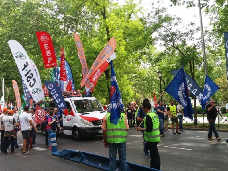 Una multitud de personas con banderas y pancartas en un evento público. La escena está ambientada en una calle con árboles y edificios al fondo. La gente parece estar participando en un desfile o manifestación, con varios vehículos y personalidades visibles.