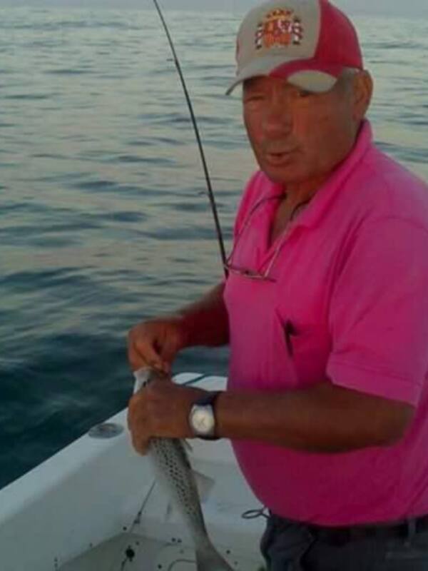 Un hombre en una camiseta rosa y una gorra blanca está pescando en el mar. La imagen muestra el mar con pequeñas ondas y el hombre está en una embarcación con una motorización visible en el fondo.