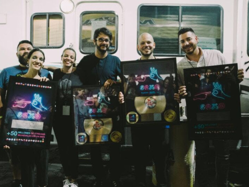 Una imagen de cinco personas sonrientes, posando con sus discos de oro y placa dorada en frente de un camión. Los discos representan ventas de más de 50 millones de copias en diferentes países, incluyendo China. La celebración parece ser para anunciar estos logros en la industria musical.