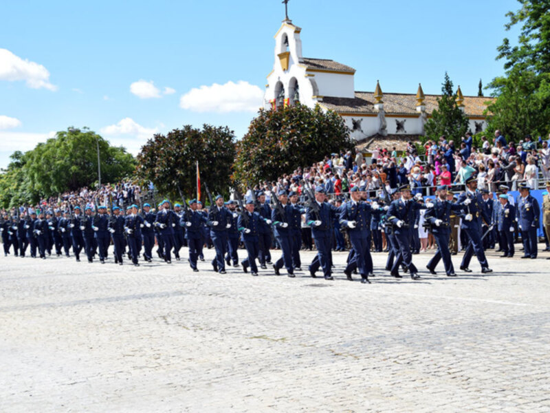 Desfile militar en una plaza con un edificio histórico al fondo. Soldados uniformizados marchan en fila, bajo un cielo azul con algunas nubes. La escena está rodeada de árboles y un público observando desde la orilla.