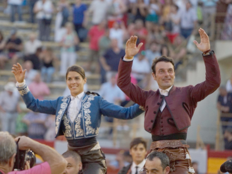 Toreros saludando a la audiencia en una corrida de toros. El torero en el centro, con traje de capote azul, y el torero en el fondo, con traje de capa roja, saludan con las manos levantadas.