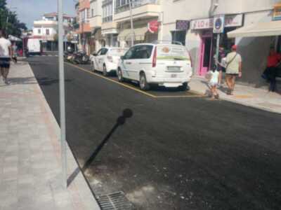 Vista de una calle con vehículos estacionados y personas caminando. Edificios comerciales a ambos lados de la calle.
