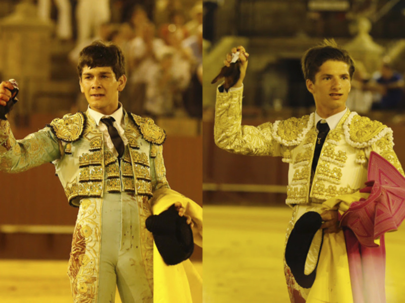 Dos toreros en su traje tradicional, uno con un chaleco dorado y otro con un chaleco blanco con detalles dorados, ambos sujetando un capote de color rojo y blanco.