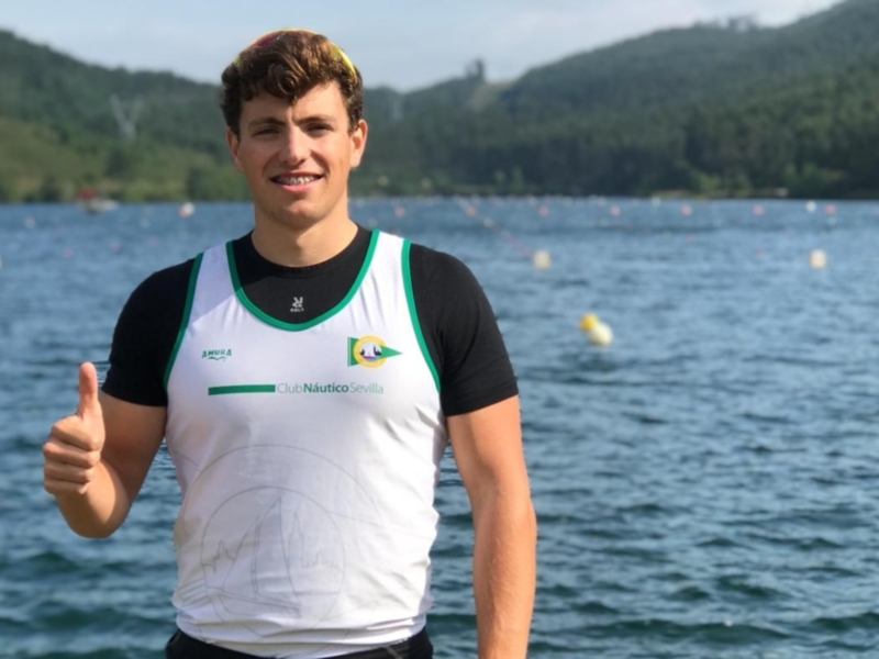 Un joven muestra un gesto de aprobación mientras está en un lago con montañas en el fondo.