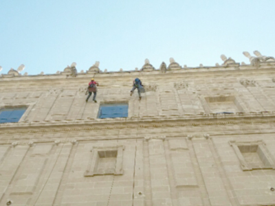 Dos personas escalan una fachada histórica de un edificio, posiblemente trabajando en la restauración o mantenimiento. La imagen muestra detalles arquitectónicos y el cielo azul claro en el fondo.