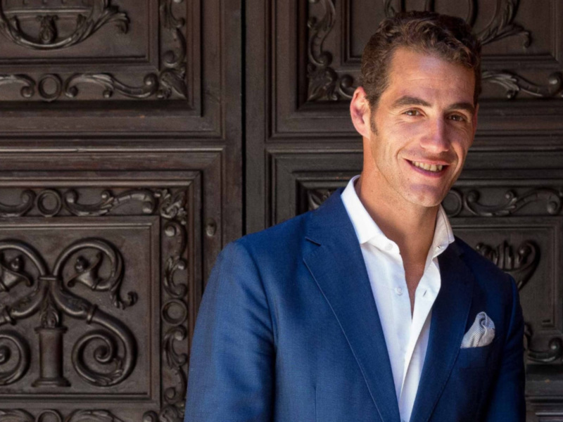 Elegante hombre en traje azul frente a una puerta de madera tallada.