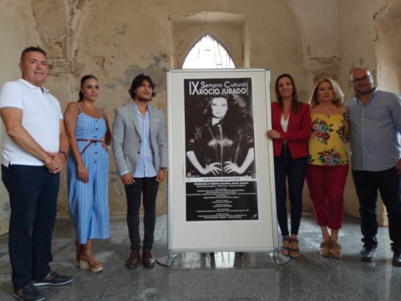 Equipo de promoción de una actividad cultural en un entorno histórico, con un cartel informativo sobre la 'IX Semana Cultural'.