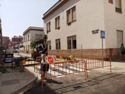 Un hombre camina por una calle con un vehículo de construcción en el fondo. Hay una señala de tráfico y una persona caminando cerca del vehículo. La imagen muestra un área urbana con edificios y una calle con señales de tráfico.