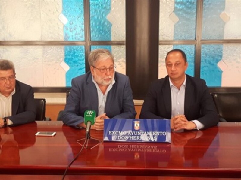Tres hombres sentados en una mesa con un micro en el centro, en una sala con una pared de cristal y un logo de la ciudad en el fondo.