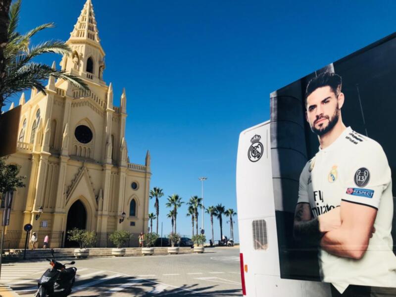 Se observa una iglesia con torres altas y detalles arquitectónicos, junto a un autobús publicitario con una imagen de un jugador de fútbol. La escena está iluminada por el sol, con árboles y una motocicleta en primer plano.